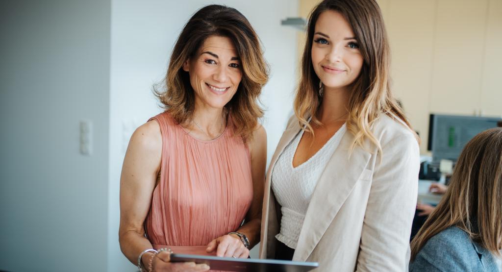 BEAUTY SOLUTIONS Team | Betreuung B2B Kunden | © BEAUTY SOLUTIONS | Martin Hofmann Fotografie Zwei Frauen stehen lächelnd in einem modernen Büro. Eine Frau hält ein Tablet, während sie sich in formeller Geschäftskleidung präsentieren. Der Raum im Hintergrund ist hell und gut beleuchtet, sorgt für eine freundliche und professionelle Atmosphäre.