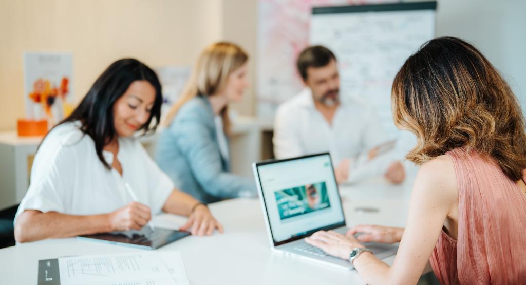 BEAUTY SOLUTIONS Teamarbeit im modernen Büro | © BEAUTY SOLUTIONS | Martin Hofmann Fotografie Geschäftsleute arbeiten konzentriert in einem modernen Büro zusammen. Eine Frau tippt auf ihrem Laptop, während andere Notizen machen und Dokumente besprechen. Ein Whiteboard ist im Hintergrund sichtbar. Die Szene vermittelt eine produktive und kollaborative Atmosphäre.