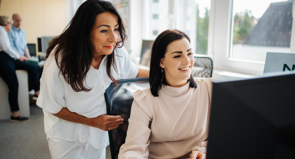 BEAUTY SOLUTIONS Team | Außendienst & Kundenservice | © BEAUTY SOLUTIONS | Martin Hofmann Fotografie Zwei Frauen arbeiten gemeinsam an einem Computer in einem modernen Büro. Die ältere Frau steht, während die jüngere sitzt und auf den Bildschirm schaut. Im Hintergrund befinden sich Fenster, durch die helles Tageslicht hereinströmt. Eine entspannte und produktive Arbeitsatmosphäre ist spürbar.