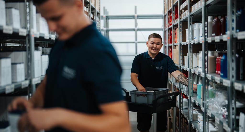 Zwei Mitarbeiter in einem Lagerhaus arbeiten effizient zwischen den Regalen. Einer von ihnen zieht einen Trolley und lächelt, während der andere konzentriert arbeitet. Die Regale sind mit verschiedenen Produkten gut gefüllt, was auf einen gut organisierten Arbeitsbereich hinweist.