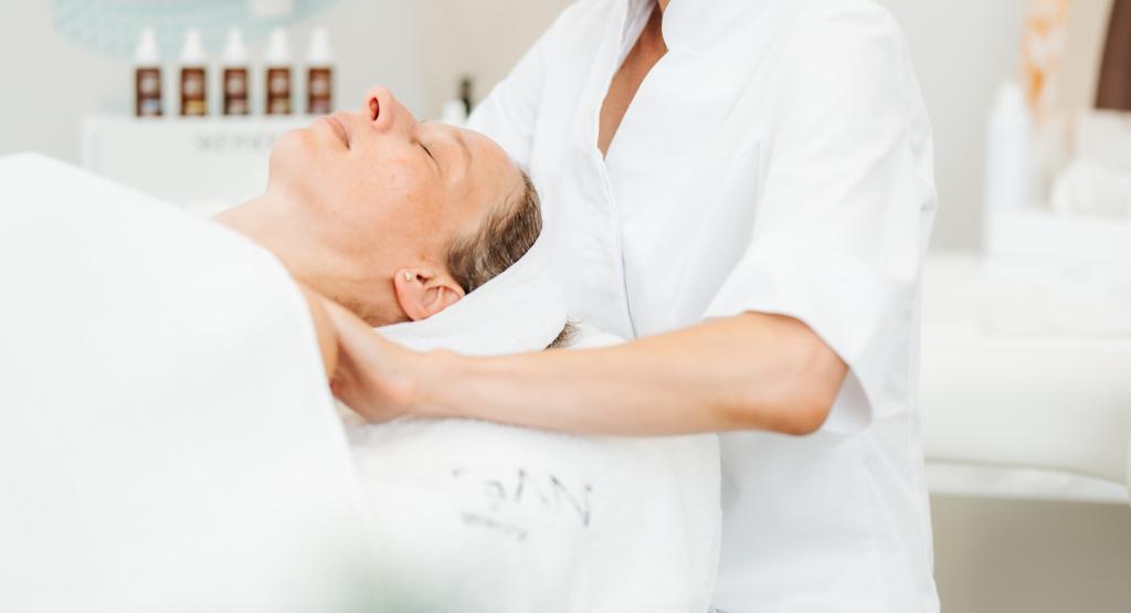 BEAUTY SOLUTIONS Training | Professionelle Gesichtsbehandlung | © BEAUTY SOLUTIONS | Martin Hofmann Fotografie Eine Kosmetikerin führt eine beruhigende Gesichtsbehandlung in einem modernen Spa durch. Die Kundin liegt entspannt auf einer weißen Liege, während die Behandlung mit Sorgfalt durchgeführt wird. Im Hintergrund sind Pflegeprodukte zu sehen, die die professionelle Umgebung ergänzen.