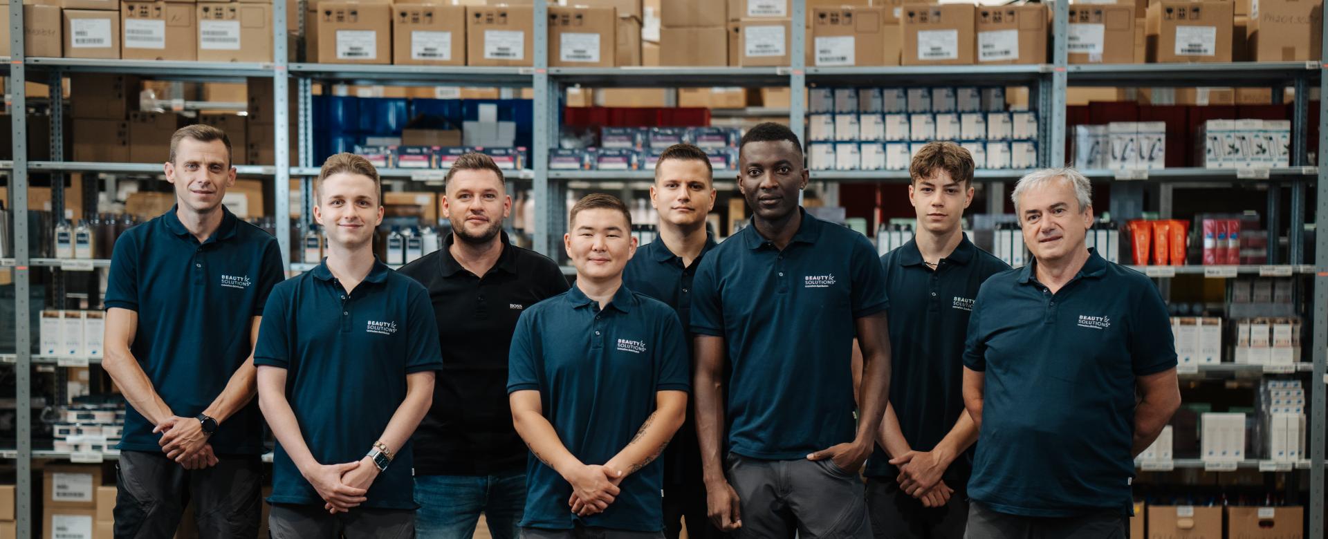 BEAUTY SOLUTIONS Team Logistik | Lager & Versand | © BEAUTY SOLUTIONS | Martin Hofmann Fotografie
Ein Teil der BEAUTY SOLUTIONS Logistik Mitarbeiter posiert in einem Lager, umgeben von Regalen und Kisten. Sie tragen einheitliche, dunkelblaue Polohemden und Arbeitsshorts, was die Teambindung verstärkt. Die Umgebung wirkt organisiert und professionell.