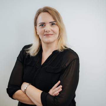 BEAUTY SOLUTIONS Team | Brand- & Office Manager | Michaela Hofgärtner | © BEAUTY SOLUTIONS | Martin Hofmann Fotografie Eine selbstbewusste Frau in schwarzer Bluse steht vor einem neutralen Hintergrund. Sie hat blonde Haare und ein zartes Lächeln, während ihre Arme verschränkt sind. Die sanfte Ausleuchtung betont den professionellen Ausdruck.