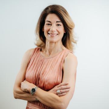 BEAUTY SOLUTIONS Team | Training Manager | Petra Juričev | © BEAUTY SOLUTIONS | Martin Hofmann Fotografie Eine lächelnde Frau in einem ärmellosen rosa Kleid posiert selbstbewusst mit verschränkten Armen vor einem neutralen Hintergrund. Ihr langes Haar fällt offen über die Schultern. Der Ausdruck im Gesicht vermittelt Freundlichkeit und Professionalität.