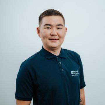 BEAUTY SOLUTIONS Team | Order Picking Specialist | Bat-Orgil Dorjkhand | © BEAUTY SOLUTIONS | Martin Hofmann Fotografie Ein Mann in einem dunkelblauen Poloshirt steht vor einem neutralen Hintergrund. Er trägt ein leichtes Lächeln und hat ein Tattoo am Arm. Die Atmosphäre ist freundlich und professionell.