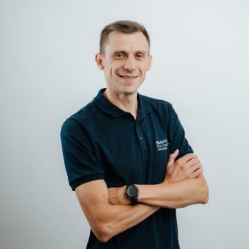 BEAUTY SOLUTIONS Team | Goods Receiving Specialist | Dimitrije Petrović | © BEAUTY SOLUTIONS | Martin Hofmann Fotografie Ein lächelnder Mann steht mit verschränkten Armen vor einem schlichten grauen Hintergrund. Er trägt eine dunkelblaue Uniform mit einem Namensschild auf der Brust. Die entspannte Haltung und das freundliche Lächeln verleihen der Aufnahme eine positive Ausstrahlung.