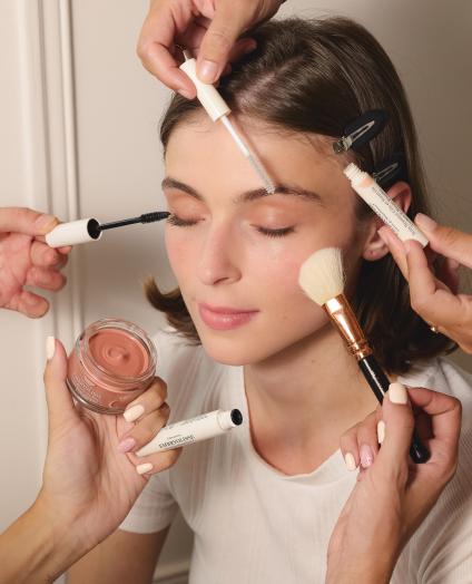 Ein Model wird von mehreren Händen mit verschiedenen Kosmetikprodukten geschminkt, darunter Pinsel und Cremes. Die Szene zeigt die Anwendung von EMBRYOLISSE-Produkten zur Erstellung eines frischen Make-up-Looks. Die neutrale Farbpalette hebt die natürliche Schönheit hervor.