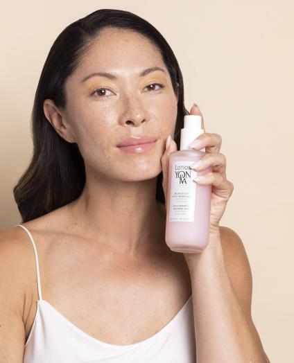 BEAUTY SOLUTIONS Kosmetikmarken | YON-KA | Lotion Hautpflege mit Feuchtigkeitsnebel | © Die Multaler Laboratorien YON-KA Eine Frau präsentiert ein Hautpflegeprodukt in einer eleganten Flasche. Sie hält es selbstbewusst in der Hand, während sie in die Kamera lächelt. Der beige Hintergrund verstärkt den Fokus auf das Produkt und die glatte Haut der Frau.