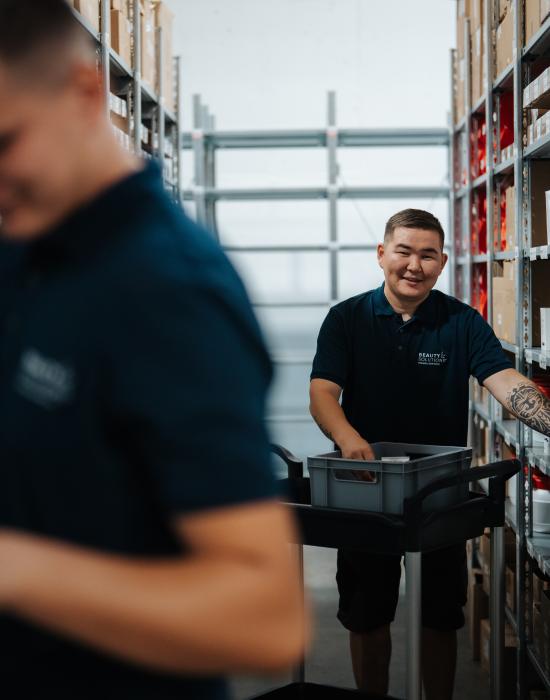 Zwei Mitarbeiter in einem Lagerhaus arbeiten effizient zwischen den Regalen. Einer von ihnen zieht einen Trolley und lächelt, während der andere konzentriert arbeitet. Die Regale sind mit verschiedenen Produkten gut gefüllt, was auf einen gut organisierten Arbeitsbereich hinweist.